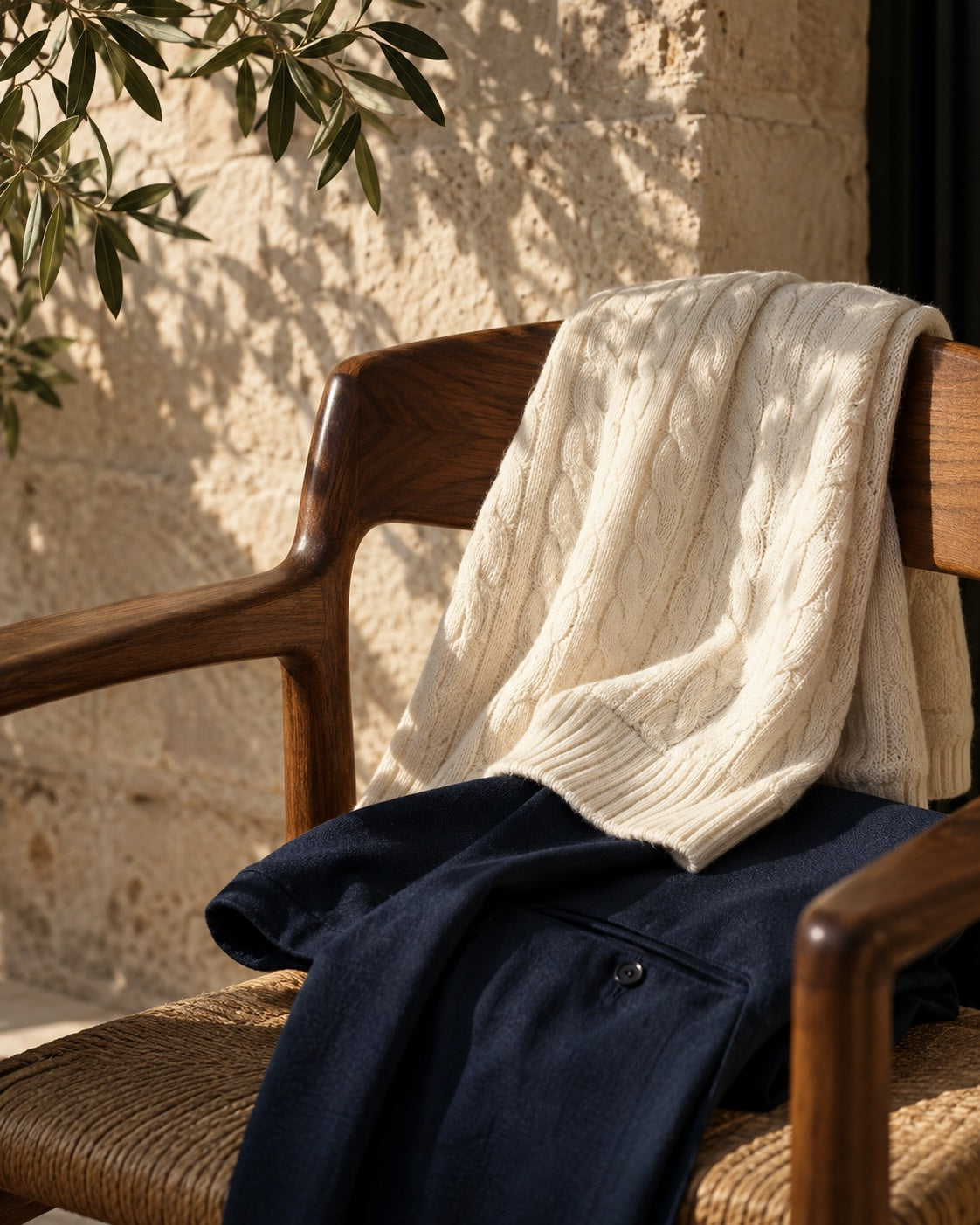 Maglia color avorio e pantaloni blu navy su una sedia in legno, con luce naturale e ombre di ulivo